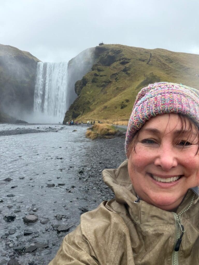 Kimberly Lorah smiling in front of Skógafoss waterfall in Iceland, wearing a colorful knitted hat and rain jacket.
