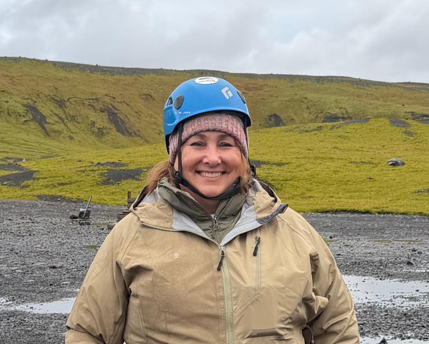 Kimberly Lorah smiling in a blue climbing helmet and beige jacket against the green hills of Iceland.